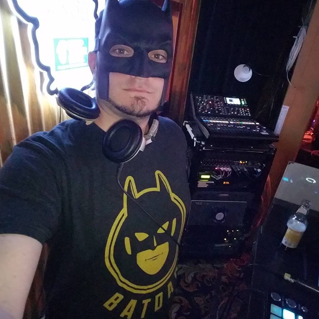 Kronik in Batman mask and headphones in the DJ booth with gear racks behind him
